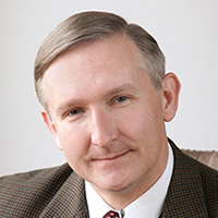 A middle-aged man wearing a tweed jacket and tie, with short hair and a mustache, poses for a professional portrait at The Hollis Law Firm.