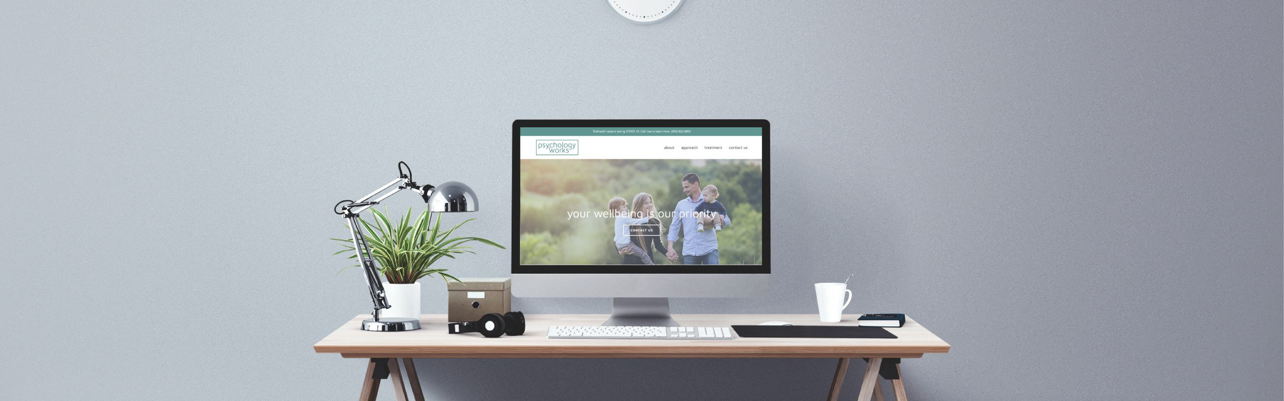 A modern workspace featuring a desk with a computer monitor displaying a website, a desk lamp, a potted plant, a small cardboard box, and a mug, against a plain gray wall.