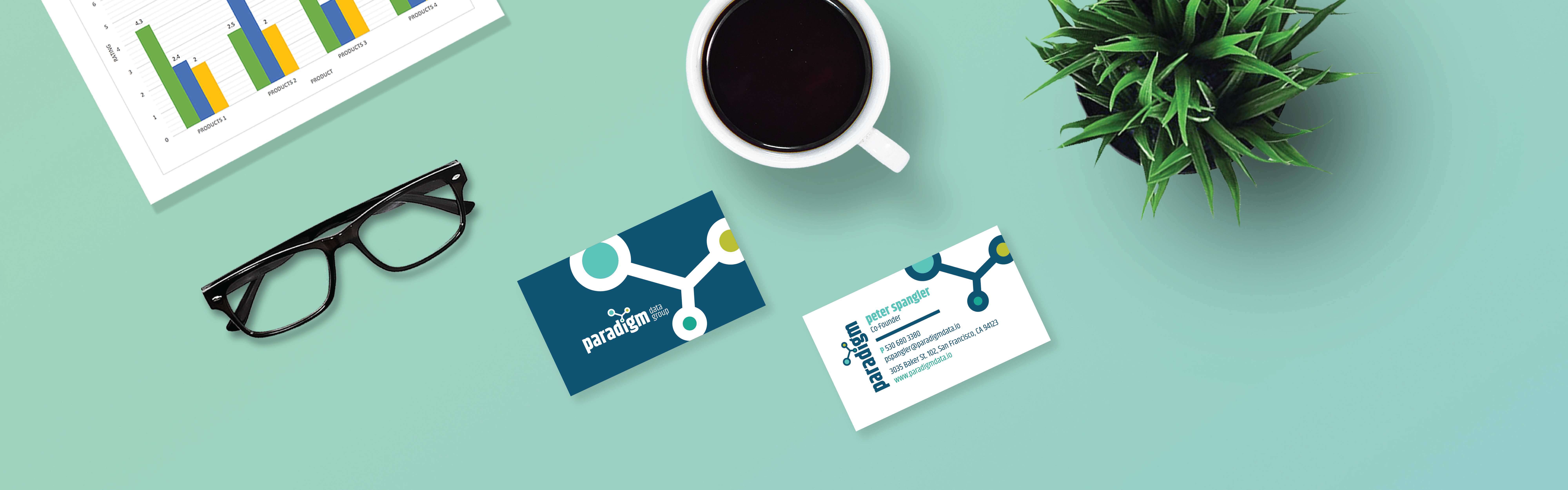 A neatly arranged desk with business cards from Paradigm Data Group, a cup of coffee, eyeglasses, a plant, and printed data charts on a turquoise background.