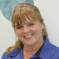A smiling woman with blonde hair wearing a blue shirt and earrings against a wall with the "Planet Kids Learning Center" blue circular pattern.