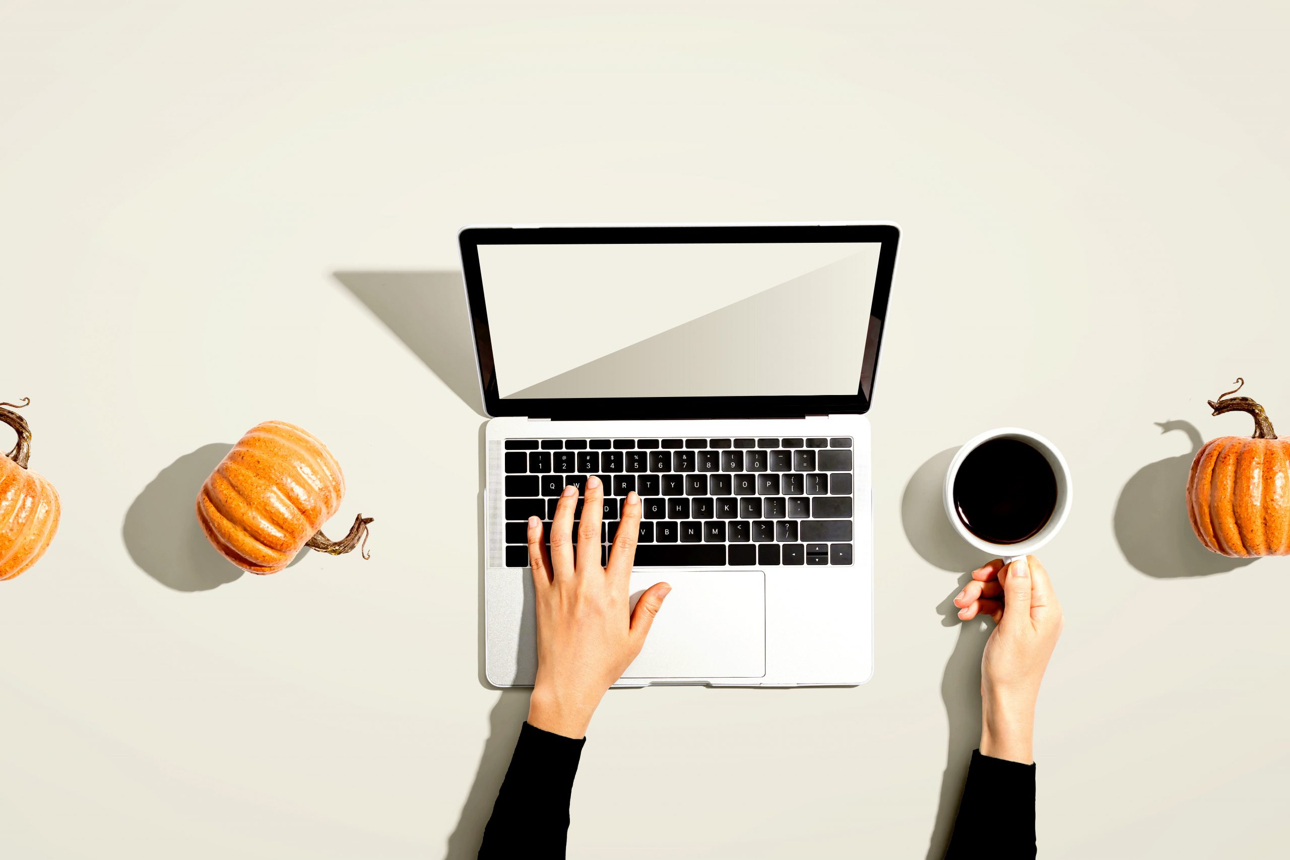 A person working on a laptop with a cup of coffee and pumpkins on the table, capturing a fall theme filled with gratitude.