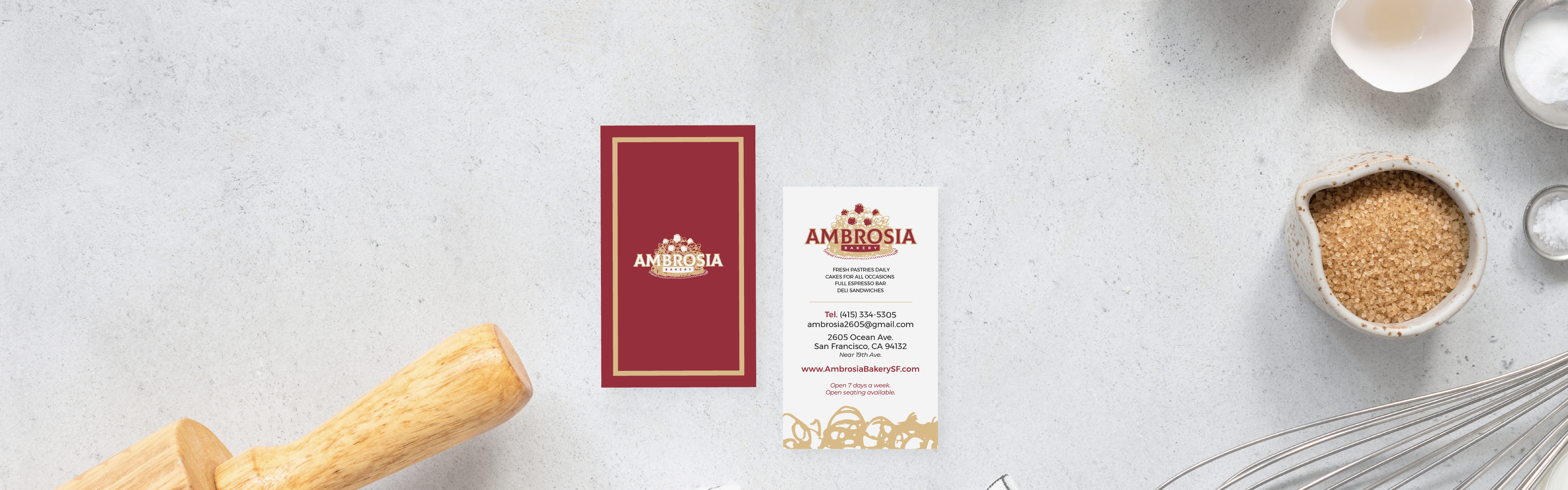A flat lay of Ambrosia Bakery's branding materials including a menu and a business card, displayed on a concrete surface alongside a rolling pin, whisk, bowl, and a small dish of brown sugar