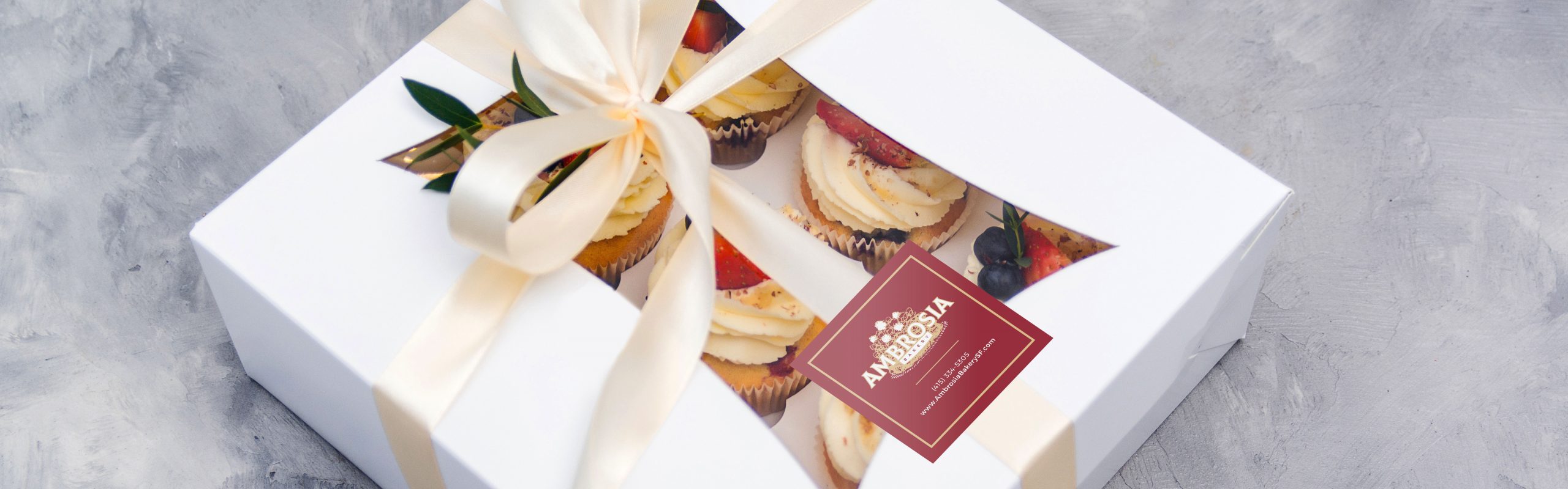 A white gift box from Ambrosia Bakery with a beige ribbon tied in a bow, containing assorted cupcakes and a tag that says "happy anniversary.