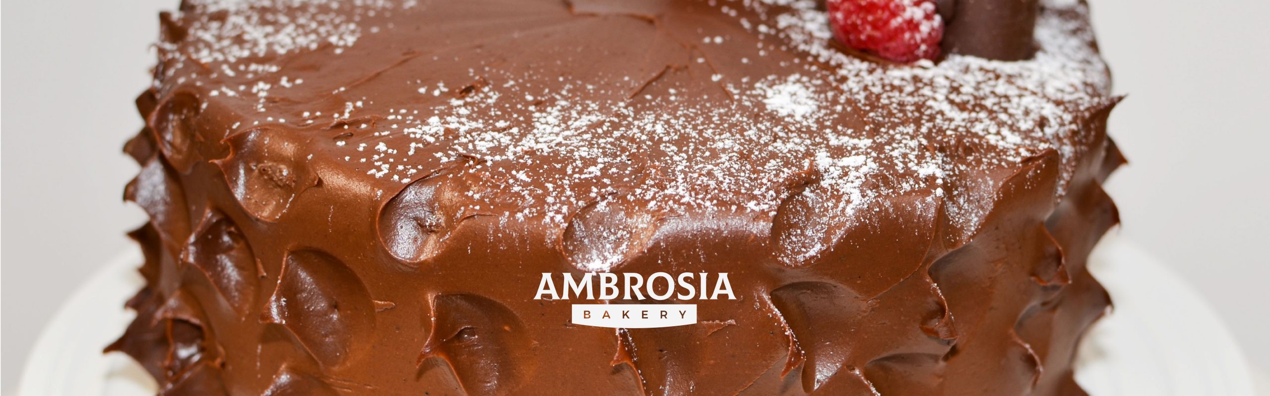 A close-up view of a chocolate frosted cake with the text "Ambrosia Bakery" on it, garnished with a dusting of powdered sugar and a single raspberry on top.