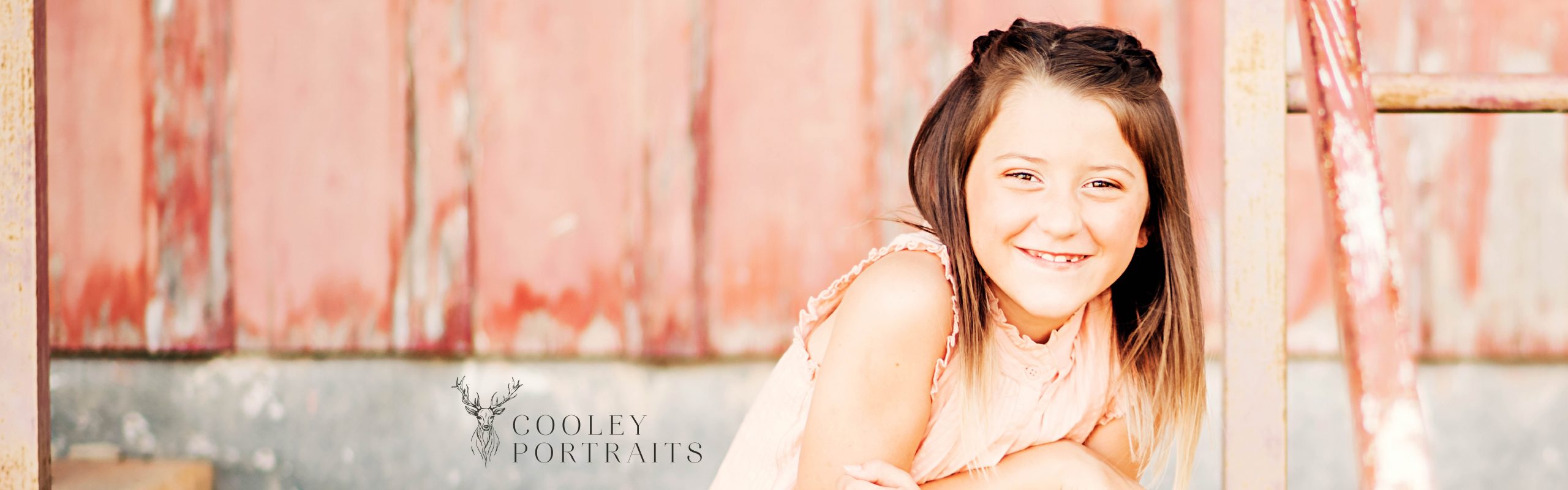 A smiling young girl leaning on a metal railing with a faded red background, captured in the unique style of Cooley Portraits.
