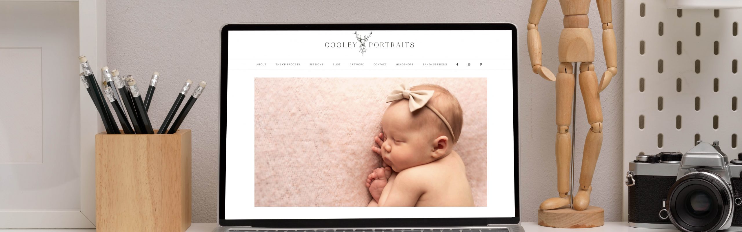 A laptop on a desk displaying a website featuring an image of a sleeping baby with a bow on its head; next to the laptop are pencils in a holder, a wooden artist's mannequin,