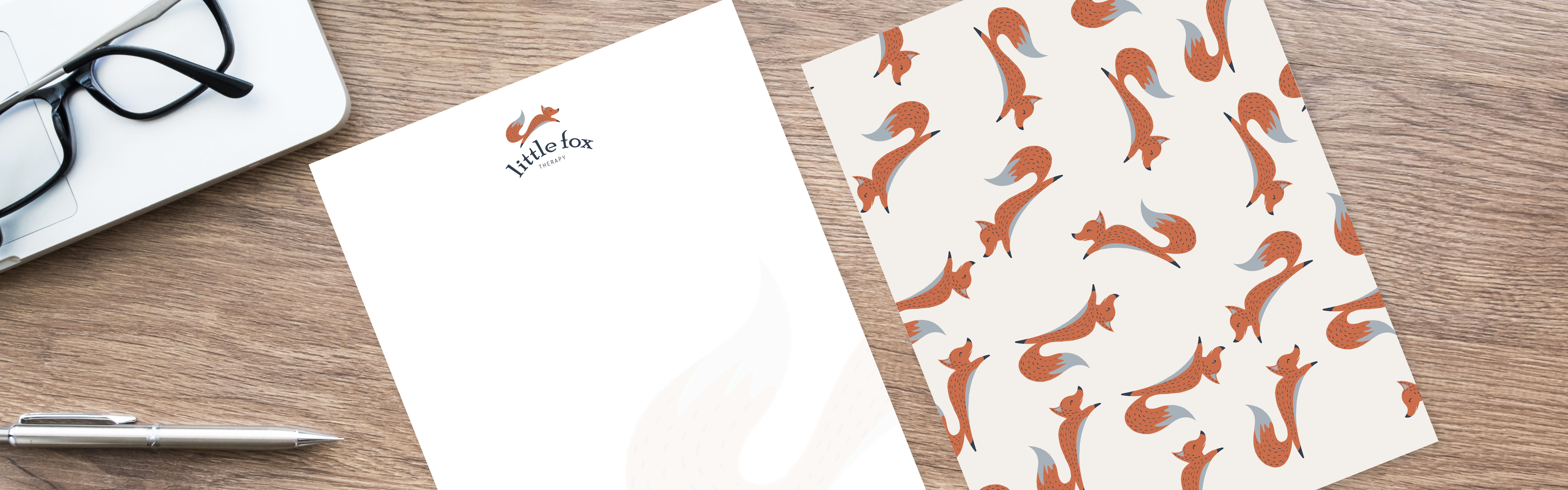 A pair of Little Fox Therapy stationery papers with fox patterns next to a silver pen and glasses on a desk.