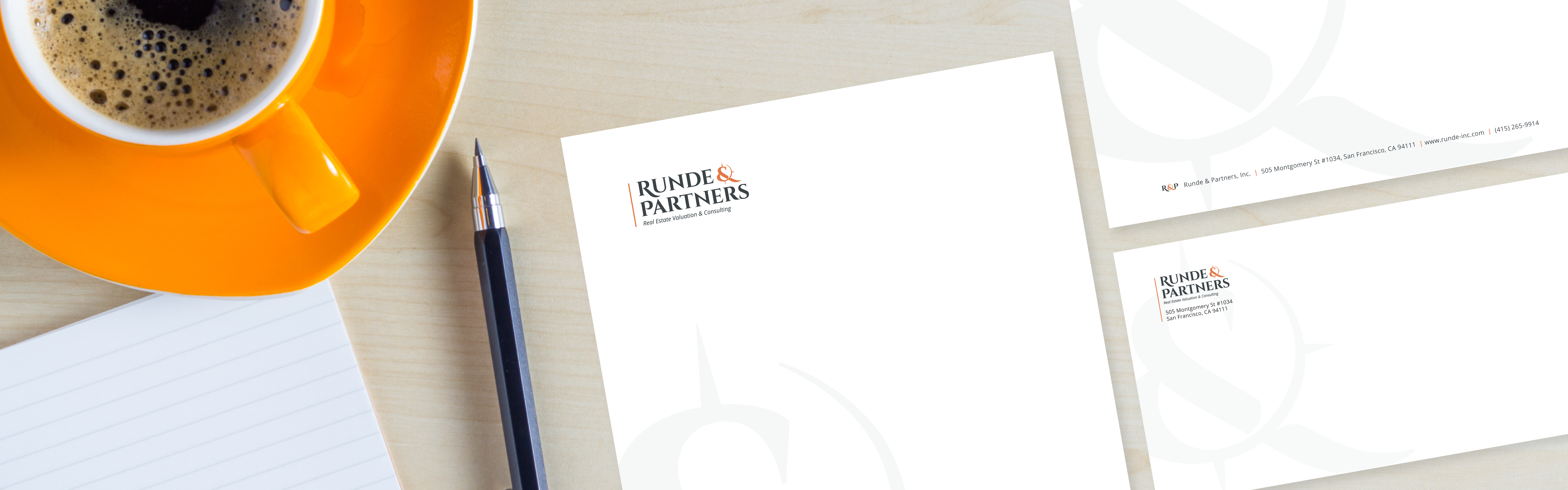 A desk setup featuring an orange coffee cup, a pen, a notepad, and letterheads for 'Runde & Partners'.