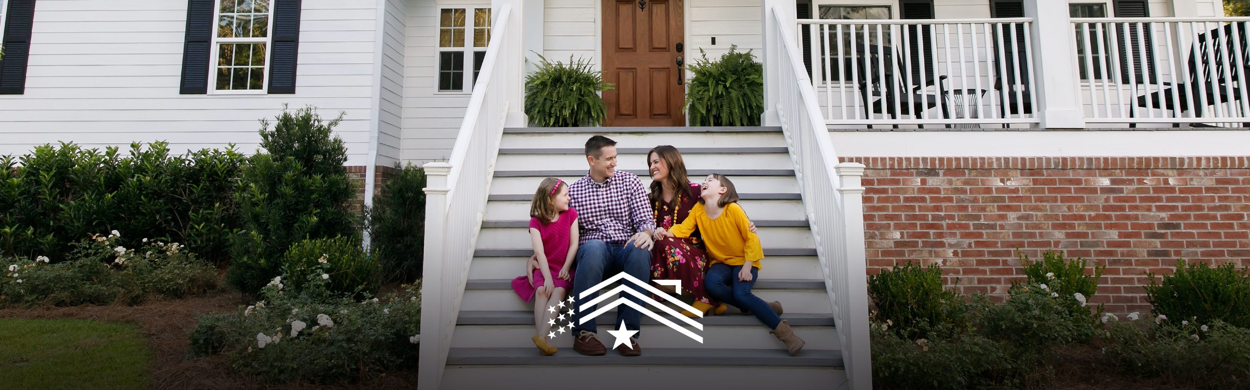 A family consisting of two adults and two children is sitting on the steps of a white house with a brown door, engaging in what appears to be a light-hearted conversation on the homefront.