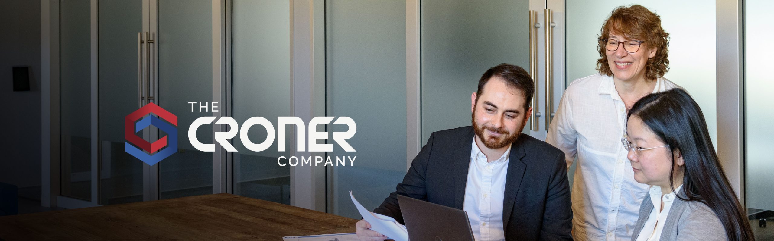 Three professionals reviewing documents together in a conference room with The Croner Company logo displayed on the wall.